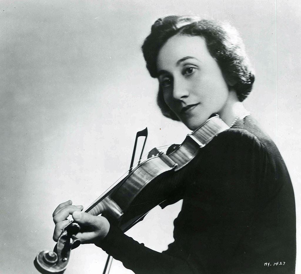 This image shows a woman playing the violin. She is wearing a black shirt and has 1940s style hair. She is holding the bow in her right hand and has her left hand on the neck of the instrument. She has a neutral expression on her face.