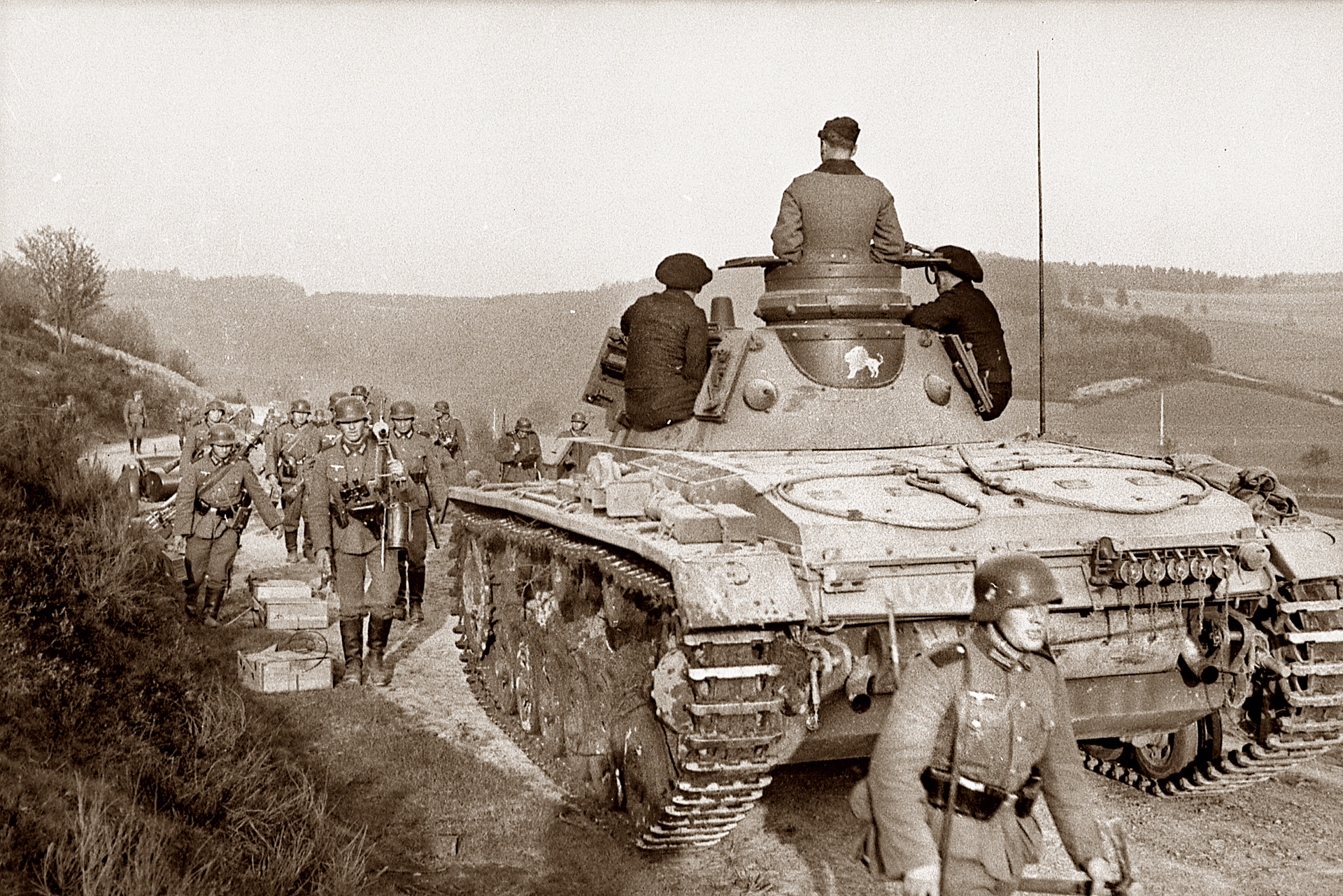 Sepia-toned historical photograph depicting a column of German infantry soldiers marching alongside a Panzer III tank, with crew members positioned on the vehicle as it travels down a rural path.